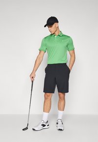Grøn polo skjorte og mørke shorts båret af en person, der holder en golfkølle. Sorte caps og hvide sneakers fuldender det sporty look.