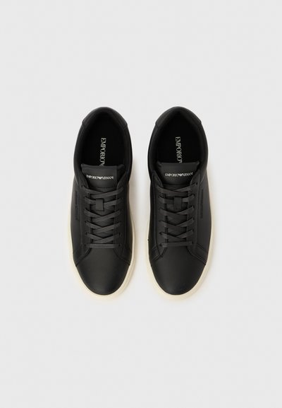 Zapatillas negras de cuero con suelas de goma blancas, que presentan punteras redondeadas, cordones planos y un logo en la lengüeta y el lateral. Textura lisa.