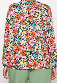 Vista posteriore di una persona che indossa una maglietta a maniche lunghe colorata con fiori arancioni, gialli, rosa e verdi, abbinata a pantaloni verde oliva.
