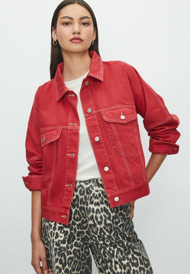 Red denim jacket with button closures, two front pockets, and white stitching. Worn over a white top and paired with leopard-print pants.