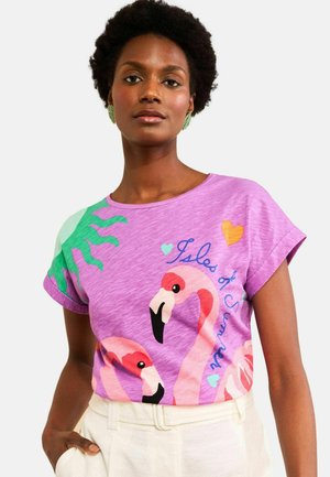 Nő lila pólóban, melyen rózsaszín flamingó rajzok és kék "Isles of Summer" felirat látható, krémszínű nadrággal párosítva, egyszínű háttér előtt áll.