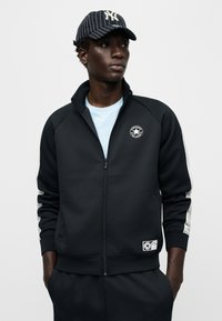 Μαύρο μπουφάν με φερμουάρ και ράγκλαν μανίκια, με στρογγυλό λογότυπο και γκρι ρ ίγες διακοσμητικά. Φορεμένο πάνω από ανοιχτό μπλε πουκάμισο.