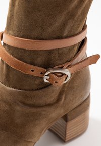 Brown suede ankle boot with a stacked wooden heel, featuring a thin tan leather strap with a silver buckle accent.