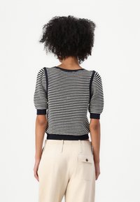 Maglione lavorato a maglia a righe blu navy e bianche con maniche corte a palloncino. Collo e orlo a coste. Abbinato a pantaloni a vita alta color crema.