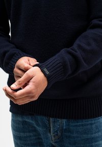 Marineblauwe gebreide trui met geribbelde boorden en onderkant. Bevat een klein etiket op de boord. Handen zijn gevouwen voor een lichte achtergrond.