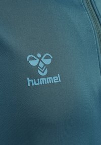 Tela de forro polar azul con una textura suave, que presenta un logotipo "hummel" en azul claro y un diseño estilizado, situado en el lado izquierdo.
