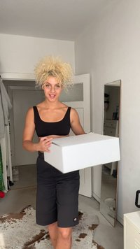 Caja rectangular blanca sostenida por una persona con cabello rubio rizado que lleva una camiseta negra y pantalones cortos. Habitación de colores claros con un espejo y muebles.