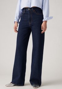 Jean taille haute bleu foncé à jambes larges avec une texture denim lisse. Doté d'une fermeture à boutons et de coutures visibles.