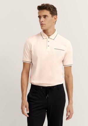 Helles rosa Polohemd mit einer Knopfleiste mit drei Knöpfen, kontrastierenden Kragenbesätzen und einer Brusttasche. Kombiniert mit schwarzen Hose mit Zugband.