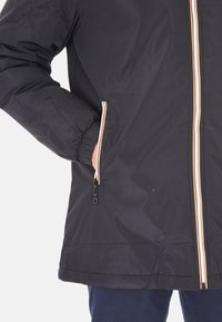 Giacca nera con polsini elastici, zip a righe multicolori e bordi delle tasche, mano inserita nella tasca con zip, indossata sopra pantaloni blu scuro.