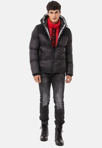 Schwarze Daunenjacke mit rotem Innenfutter, ausgestattet mit einer Kapuze, Reißverschluss und Logodetails. Kombiniert mit dunklen Jeans und schwarzen Stiefeln.