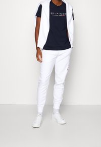 Námořnické tričko s logem "HILFIGER NEW YORK", bílá mikina přehozená přes ramena, bílé jogger kalhoty a bílé tenisky.