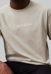 Beige bomulds t-shirt med rund hals, prydet med hvidt præget "Calvin Klein" logo på fronten. Glat tekstur, korte ærmer.