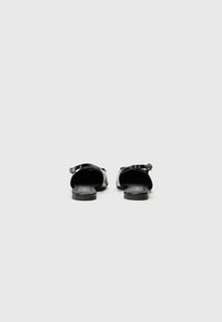 Black heeled sandals featuring a glossy finish, open back, adjustable buckle strap, and flat sole. Smooth texture with a minimalist design.