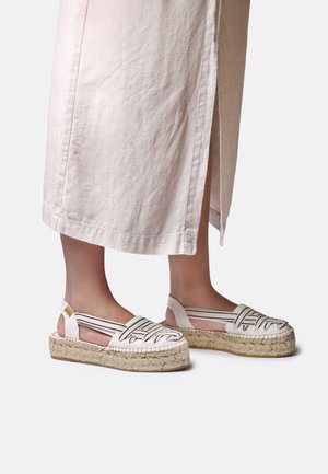 Nohy v bielych espadrilových sandáloch s tkanými podrážkami a elastickými pásmi s čiernymi pruhmi, doplnené béžovou sukňou s bočným rozparkom.