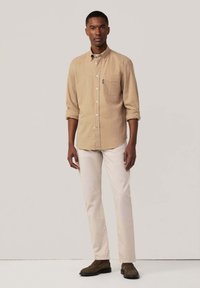 Camisa beige de botón con mangas largas y bolsillo en el pecho, combinada con pantalones crema claros y zapatos marrón oscuro. Diseño simple y limpio.