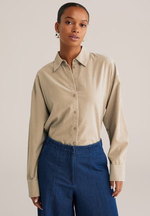 Woman with silver hoop earrings wearing a beige button-up shirt tucked into high-waisted dark blue pants against a plain background.