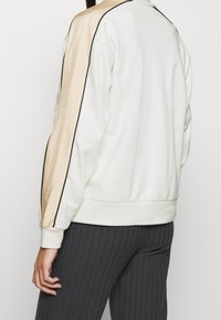 Vit sweatshirt med guldiga satinliknande sidostråkar, svarta pipningar, ribbade ärmslut och nederkant. Slät textur, avslappnad passform, minimalistisk design.