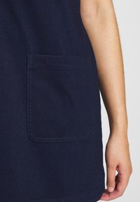 Oikea käsi tummansinemmän vaatteen vieressä, jossa on suorakulmainen läpässä tasku edessä, lähikuva tasaisen taustan edessä.