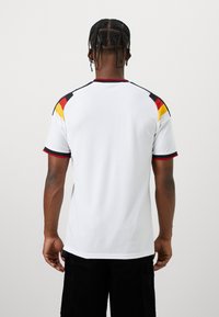 Homme aux cheveux tressés portant un maillot de sport blanc avec des accents rouges, jaunes et noirs, vu de dos sur un fond uni.