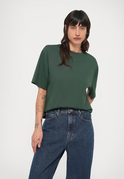 Σκούρο πράσινο κοντομάνικο μπλουζάκι με άνετη εφαρμογή, στρογγυλή λαιμόκοψη και λεία υφή, συνδυασμένο με ψηλόμεσα μπλε τζιν.