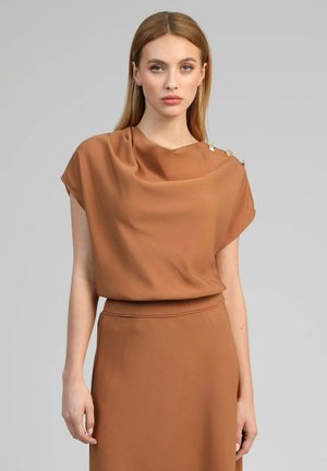 Femme aux cheveux raides châtain clair portant une robe marron sans manches avec des boutons dorés sur l'épaule, debout devant un fond gris uni.