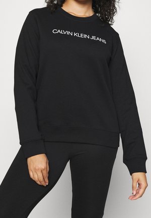 Kobieta nosząca czarną bluzę z długim rękawem z białym logo "Calvin Klein Jeans" na piersi, zestawioną z czarnymi spodniami.