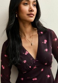 Abito floreale scuro a maniche lunghe, con motivi di rose rosa, scollatura a V e rifiniture in pizzo. Collana dorata con pendente a forma di iniziale.