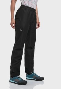 Schwarz wasserfeste Hose mit elastischem Bund, Reißverschlusstaschen und reflektierendem Logo. Kombiniert mit blauen und grauen Wanderschuhen.