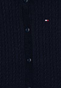 Marineblå strikket cardigan med kabelmønster, knappelukking og liten rød, hvit og blå logo detalj. Myk tekstur og ribbede kanter.