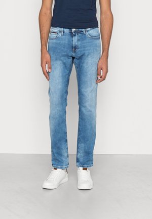 Mężczyzna w jasnoniebieskich dżinsach o prostych nogawkach, białych tenisówkach i granatowej koszulce, stojący na tle jednolitego jasnego tła.