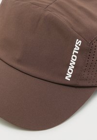 Καφέ υφασμάτινο καπέλο με κυρτή γείσο και λευκό κείμενο "SALOMON" στην πλευρά, με μικρές τρύπες αερισμού κοντά στο κείμενο.