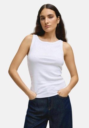 Femeie cu păr lung și închis la culoare, purtând un top alb fără mâneci și blugi bleumarin, stând cu mâinile în buzunare pe un fundal simplu.