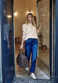 Jasnobeżowa bluzka z długim rękawem i guzikami, niebieskie jeansy o szerokich nogawkach, trzymająca brązową skórzaną torbę podróżną, białe trampki, wnętrze pomieszczenia.