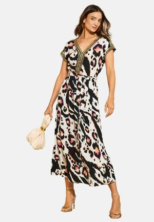 Frau trägt ein knielanges Kleid mit animalischem Muster in Schwarz, Weiß und Pink mit goldener Verzierung, hält eine beige Clutch und trägt goldene Sandaletten mit hohen Absätzen.