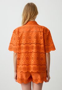 Oranje blouset met eyelet-details, korte mouwen en een geschulpte zoom, voorzien van intricieuze bloemmotieven, gecombineerd met bijpassende shorts.
