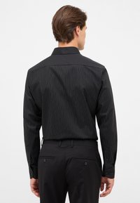 Mann in einem schwarzen Streifenhemd mit langen Ärmeln, das in schwarze Hosen gesteckt ist, von hinten gesehen vor einem schlichten weißen Hintergrund.