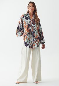 Camisa colorida y estampada con un corte relajado, que presenta mangas oversized y un diseño de botones, combinada con pantalones de pierna ancha en color crema.