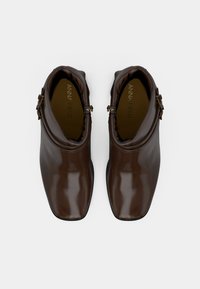 Botas de cuero marrón hasta el tobillo con un acabado brillante, puntera afilada y detalle de hebilla lateral. Interior forrado con nombre de marca en relieve.