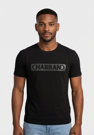 Hombre con cabello corto y barba, vistiendo una camiseta negra con el texto "CHABRAND" y jeans azul claro, de pie frente a un fondo blanco liso.