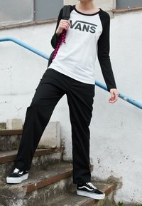 Vans langærmet skjorte i hvid og sort, sorte bukser og sorte sneakers med hvide accenter, stående på trin nær et blåt gelænder.