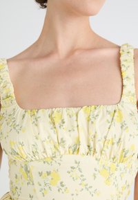 Kvetinové midi šaty zo svetložltého materiálu. Majú štvorcový výstrih a nariasené ramienka. Po celej ploche sú malé žlté a zelené kvety.