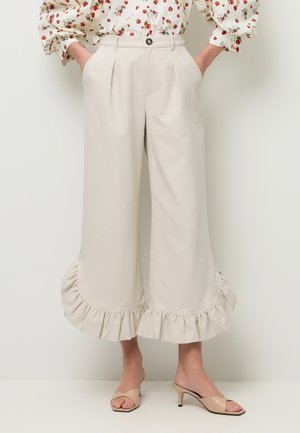 Beżowe krótkie spodnie o szerokich nogawkach z falbanami na zakończeniach, plisami z przodu i bocznymi kieszeniami, noszone z beżowymi odkrytymi mulesami na niskim obcasie.