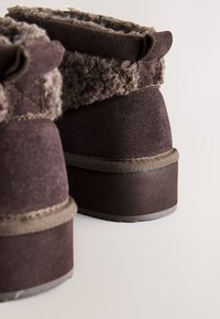 Paire de bottes d'hiver en daim brun foncé avec semelles épaisses en caoutchouc et bordure en laine duveteuse autour de la cheville.