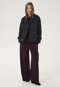 Giacca nera in cotone doppiopetto con grandi bottoni, abbinata a pantaloni morbidi bordeaux. L'outfit presenta un colletto e tasche laterali.