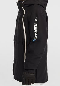 Personne portant une veste noire O'Neill avec des rayures verticales blanches et un logo sur la manche, associée à des gants noirs Dakine, vue de profil.