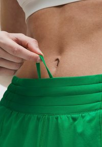 Pantalones cortos deportivos verdes con una cintura texturizada. Una mano tira de un cordón a juego, destacando el diseño elástico y la cintura ajustada.