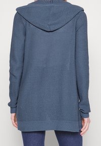 Lange, blauwe cardigan met een capuchon, voorzien van een textuurgebreid patroon en geribbelde manchetten en zoom. De achterkant is zichtbaar, waarbij de lengte en pasvorm worden benadrukt.