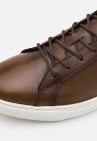 Baskets en cuir marron avec une finition lisse, bout arrondi et semelle en caoutchouc blanc. Elles présentent des lacets et des détails de couture marron foncé.