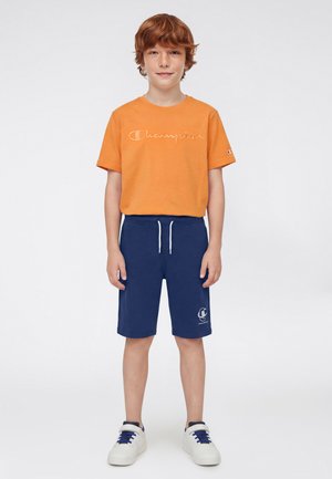 Ragazzo dai capelli rossi in piedi, indossa una maglietta arancione Champion, pantaloncini blu navy con coulisse e scarpe da ginnastica bianche con lacci blu.